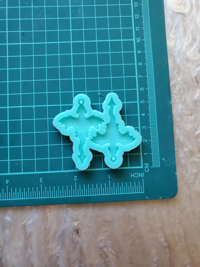 May include: Two teal blue silicone molds of a dragon with wings outstretched and a tail that ends in a point. The molds are on a green cutting mat with inch and centimeter markings.