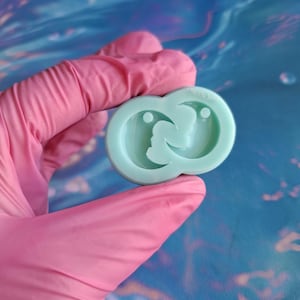 Op de afbeelding: Een lichtgroene siliconen mal met twee ronde indrukken van een halve maan met een kleine cirkel in het midden van elke maan.