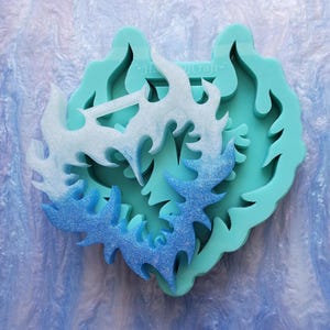 Könnte beinhalten: Eine tealblaue Silikonform in Herzform. Die Form hat ein Design aus drei überlappenden Flammenformen mit einem Farbverlauf von Weiß zu Blau.