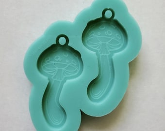 May include: A light teal silicone mold featuring two mushroom designs. Each mushroom has a detailed cap with small circles and a long stem. The mold is designed for crafting and has a small loop at the top of each mushroom for hanging.