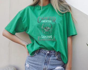 May include: A green t-shirt featuring the Cancer zodiac sign. The design includes the words "Cancer Creative Loving Emotional" in a stylized font, along with a crab graphic. The shirt is paired with denim shorts.