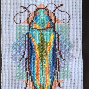 Puede incluir: Un diseño de punto de cruz de un insecto estilizado, con un cuerpo central turquesa con detalles en naranja y verde. El diseño incluye detalles en marrón, melocotón y azul, sobre un fondo de tela blanca. El insecto tiene una forma geométrica.