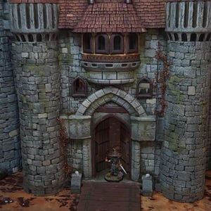 May include: A detailed miniature castle model with stone walls, brown tiled roofs, and two tall towers. A wooden drawbridge leads to the arched doorway, where a miniature figure stands. The castle is set on a brown surface.
