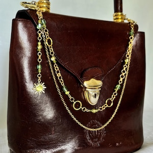 以下が含まれることがあります： ゴールドトーンの金属製クラスプと装飾的なゴールドチェーンが付いたダークブラウンのレザーハンドバッグ。アクセサリーには小さな緑色のビーズと太陽の形をしたチャームが含まれており、トップハンドルからバッグの前面に垂れ下がっています。