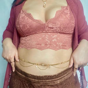 May include: A gold-colored body chain with delicate leaf and crystal charms. The chain is worn over a brown lace top and a matching skirt. A pink lace bralette and a pink cardigan are also visible.