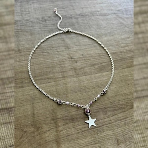 Könnte beinhalten: Silberfarbene Halskette mit filigraner Kette und Sternanhänger. Die Halskette hat kleine, runde, lila Perlen. Der Sternanhänger hat eine einfache, fünfzackige Form. Die Halskette wird auf einer Holzoberfläche präsentiert.
