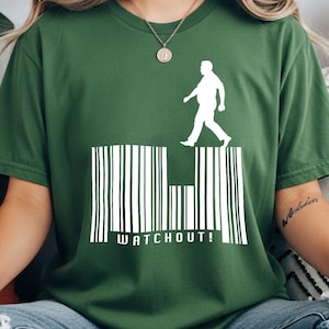 Puede incluir: Camiseta verde con un gráfico blanco de una persona caminando sobre un diseño de código de barras. La palabra "WATCHOUT!" está impresa debajo. La camiseta es de un material suave.