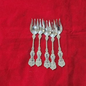 May include: Five silver forks with ornate handles are arranged on a red background. Each fork has four tines and a decorative handle with floral and scrollwork patterns. The forks are likely for serving appetizers or desserts.