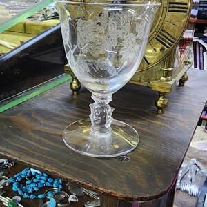 May include: A clear glass goblet with an etched floral design sits on a wooden surface next to a brass clock with Roman numerals. The clock has a decorative design and is in a vintage style.