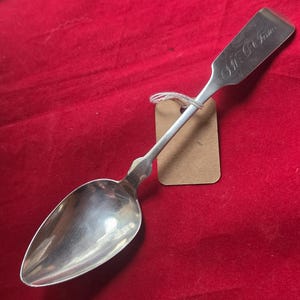 May include: A silver spoon with a long handle and a rounded bowl. The handle has an inscription and a decorative design near the bowl. A small brown tag is attached to the spoon with twine. The spoon is set against a red background.