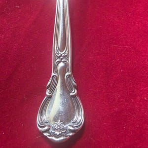 May include: Close-up of a silver fork with an ornate, detailed design. The fork's handle features a decorative pattern with curved lines and floral accents. The tines are visible at the top. The background is a solid red color.