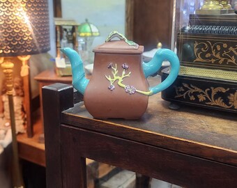 May include: A brown teapot with a blue handle and lid, decorated with a floral design. The teapot is sitting on a wooden shelf with other items.