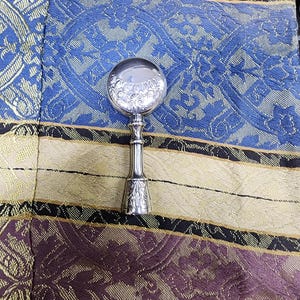 May include: A silver-colored magnifying glass with a round, clear lens and a decorative handle. The lens has a detailed floral design. The handle has a flared base with ornate carvings. The magnifying glass is set against a patterned fabric.