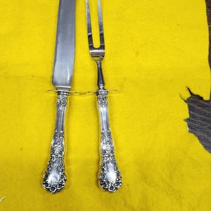 May include: A silver carving knife and fork set. The knife has a long, curved blade and the fork has two tines. Both pieces have ornate, floral-patterned handles and a decorative ring near the blade/tines.