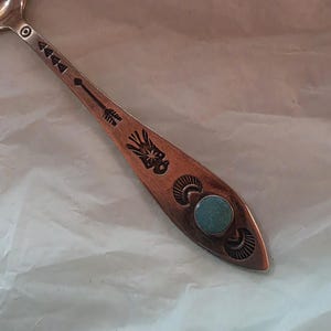 May include: A silver-toned metal spoon featuring an elongated handle decorated with stamped arrow, star, and crescent moon motifs. A small, round, light blue turquoise-colored stone is inlaid near the tip of the handle, adding a focal point.