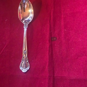 May include: A silver spoon with an ornate handle and a rounded bowl, set against a deep red background. The spoon's design features detailed patterns and curves, reflecting light. A classic piece of silverware.