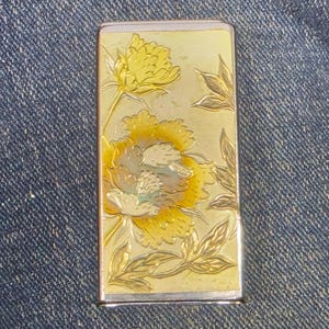 May include: A rectangular metal lighter case featuring an embossed floral design with yellow and gold tones. The surface displays detailed engravings of two blooming flowers and leaves, framed by a polished silver-colored metallic border.