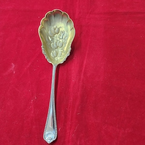 May include: A silver serving spoon with an ornate design featuring a cluster of berries. The spoon has a beaded handle and a decorative floral design at the end.