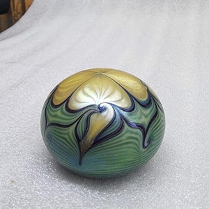May include: A decorative glass paperweight with a swirling pattern. The paperweight features a blend of colors, including gold, green, and blue. The design has a floral or abstract motif, with curved lines and a smooth, rounded shape.