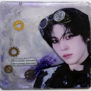 May include: A square resin coaster featuring a portrait of a person with dark hair wearing steampunk goggles. The background is a swirling mix of white and purple glitter. Four small gold-toned metal gears and the text We're pumpin' Adrenaline.