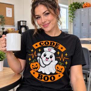 May include: A black t-shirt featuring a graphic of a white ghost dressed as a nurse with a stethoscope, flanked by two orange jack-o'-lanterns. The design includes the text CODE BOO, yellow stars, and an orange heartbeat line.