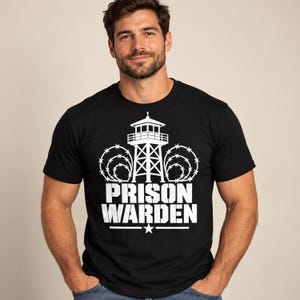 Può includere: Una t-shirt nera a maniche corte con una stampa grafica bianca raffigurante una torre di guardia circondata da filo spinato. Sotto la torre, il testo PRISON WARDEN è stampato in grassetto bianco, con una piccola stella centrata sotto il testo.