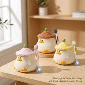 Op de afbeelding: Drie handgemaakte keramische eendenmokken met houten deksels en decoratieve lepels. De mokken zijn wit met gele snavels en poten. Eén mok heeft een roze deksel, één heeft een bruin deksel en één heeft een gele deksel. Elke mok heeft een bijpassende lepel.