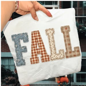 May include: A white t-shirt featuring the word FALL in large block letters. Each letter has a different pattern: the F is blue with white flowers, the A is orange and white gingham, the L is cream with floral print, and the L is tan plaid with pumpkins.
