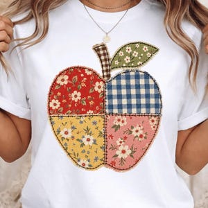 May include: A white t-shirt featuring a large apple graphic composed of four distinct fabric patches. The apple design includes red floral, blue gingham, yellow floral, and pink floral patterns, with a brown and white checkered stem and a green leaf.