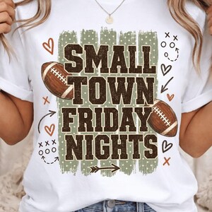 May include: A white t-shirt featuring the text "SMALL TOWN FRIDAY NIGHTS" in dark brown block letters over a sage green polka dot brushstroke background. The design includes two brown footballs, orange hearts, and white play diagrams.