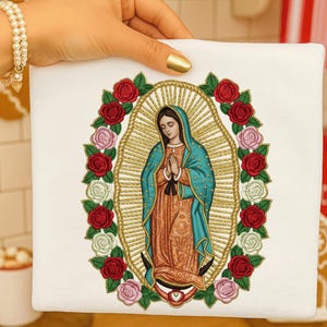 May include: A white square fabric with an embroidered image of the Virgin Mary, encircled by a gold halo and a wreath of red, pink, and white roses. The Virgin Mary is shown with hands clasped in prayer, wearing a teal cloak and orange robe.