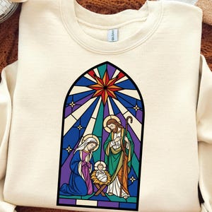 May include: Cream-colored sweatshirt featuring a stained-glass design depicting the Nativity scene. The graphic includes Mary, Joseph, and baby Jesus, with a starburst at the top. The design is framed in black.