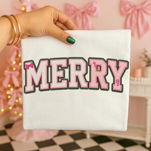 Op de afbeelding: Wit t-shirt met het woord "MERRY" in roze, omlijnd met groen. De letters hebben decoratieve elementen zoals een strik, kerstboom en zuurstokken. Het shirt wordt vastgehouden door een hand met gouden armbanden en groene glitter nagellak.