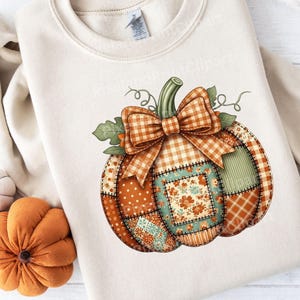 Op de afbeelding: Crèmekleurige sweatshirt met een groot, kleurrijk pompoenontwerp. De pompoen is gemaakt van patchworkstof met verschillende patronen, waaronder stippen, ruiten en bloemenprints. Een geruite strik staat bovenop de pompoen, met groene bladeren en ranken.