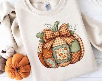 May include: Cream-colored sweatshirt featuring a large, colorful pumpkin design. The pumpkin is made of patchwork fabric with various patterns, including polka dots, plaid, and floral prints. A plaid bow sits atop the pumpkin, with green leaves and vines.