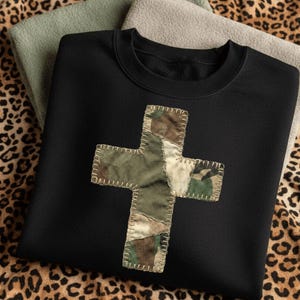 May include: Black crewneck sweatshirt featuring a patchwork cross design in camouflage fabric. The cross is stitched onto the front of the sweatshirt. The sweatshirt is displayed on a leopard print background with two folded sweatshirts.