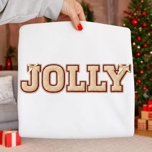 May include: White square pillow with the word "JOLLY" embroidered in tan and brown, with small bow accents. The pillow is held by a person with red nail polish. A Christmas tree is visible in the background.