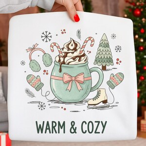 Puede incluir: Sudadera blanca con un diseño de invierno. El diseño incluye una taza verde azulado con crema batida y bastones de caramelo, mitones, orejeras, un árbol de Navidad, copos de nieve y el texto "WARM & COZY".