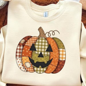 May include: Cream-colored sweatshirt featuring a patchwork pumpkin design. The pumpkin has a smiling jack-o'-lantern face and is composed of various patterned fabrics in shades of orange, brown, green, and yellow. A perfect fall or Halloween themed item.