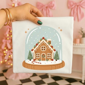 Peut inclure: Un tissu carré blanc avec un motif sur le thème de Noël. Le motif représente une maison en pain d'épices à l'intérieur d'une boule à neige, avec un fond bleu clair et de la neige qui tombe. La maison en pain d'épices a des murs bruns, du glaçage blanc et des décorations de bonbons colorées.