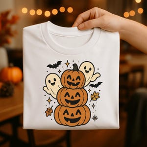 Könnte beinhalten: Weißes T-Shirt mit einem Halloween-Motiv. Das Design zeigt einen Stapel Kürbisse mit lächelnden Gesichtern, zwei Geister und Fledermäuse. Enthält Sterne und Herbstblätter.