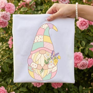 Könnte beinhalten: Ein weißes T-Shirt mit einem farbenfrohen Zwergen-Design. Der Zwerg hat einen Patchwork-Hut in Pastellfarben, einen langen weißen Bart und hält einen Blumenstrauß. Das Design ist vor einem Hintergrund aus rosa Rosen platziert.