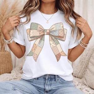 May include: White t-shirt featuring a large, patchwork bow design in pastel colors. The bow has a variety of patterns, including floral and gingham, in shades of pink, green, and beige. The shirt is a casual, everyday wear item.