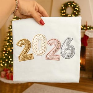 Może przedstawiać: Biały materiał z haftowanymi cyframi "2026" w kolorach złotym, kremowym, różowym i srebrnym. Cyfry mają wygląd cekinów. W tle widoczna jest choinka i kominek.