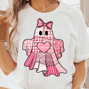 May include: White t-shirt with a pink ghost design. The ghost is made of patchwork fabric in various pink patterns, with a pink bow, heart, and black eyes. The design is centered on the shirt.
