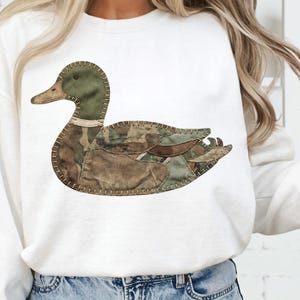 May include: White sweatshirt with a duck applique. The duck design is crafted from camouflage fabrics in green, brown, and tan, stitched together. The duck has a green head and a brown beak.