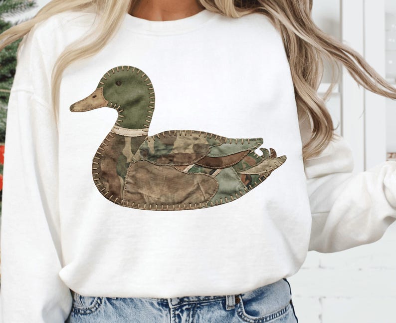 May include: White sweatshirt with a duck applique. The duck design is crafted from camouflage fabrics in green, brown, and tan, stitched together. The duck has a green head and a brown beak.