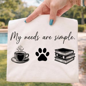 May include: Cream-colored tote bag with the phrase "My needs are simple." printed in black script. The design includes a coffee cup, a paw print, and a stack of books, all in black.