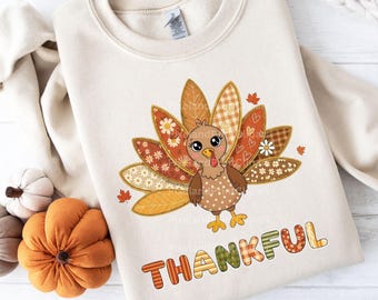 Puede incluir: Sudadera de color crema con un diseño de pavo de dibujos animados. El pavo tiene plumas de retazos y ojos grandes. La palabra "THANKFUL" está impresa debajo en letras coloridas y estampadas. Prenda de vestir con temática otoñal.