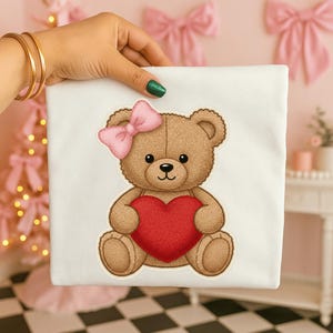 Può includere: Una borsa tote bianca con un'illustrazione di un orsacchiotto marrone. L'orso ha un fiocco rosa e tiene un cuore rosso. Lo sfondo è rosa tenue, con fiocchi decorativi e un tavolino.